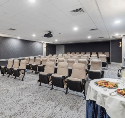 An empty theater-style room with rows of seats, a projector on the ceiling, and a table with food at the front.