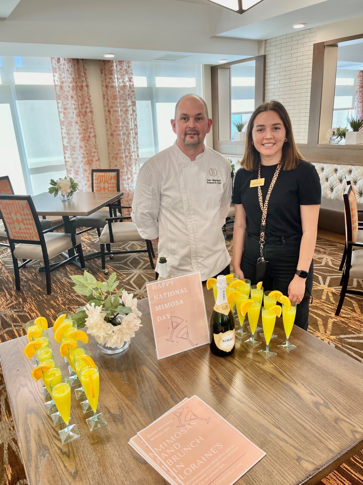 A chef and a staff member stand by a table set with champagne glasses, ready for an event.
