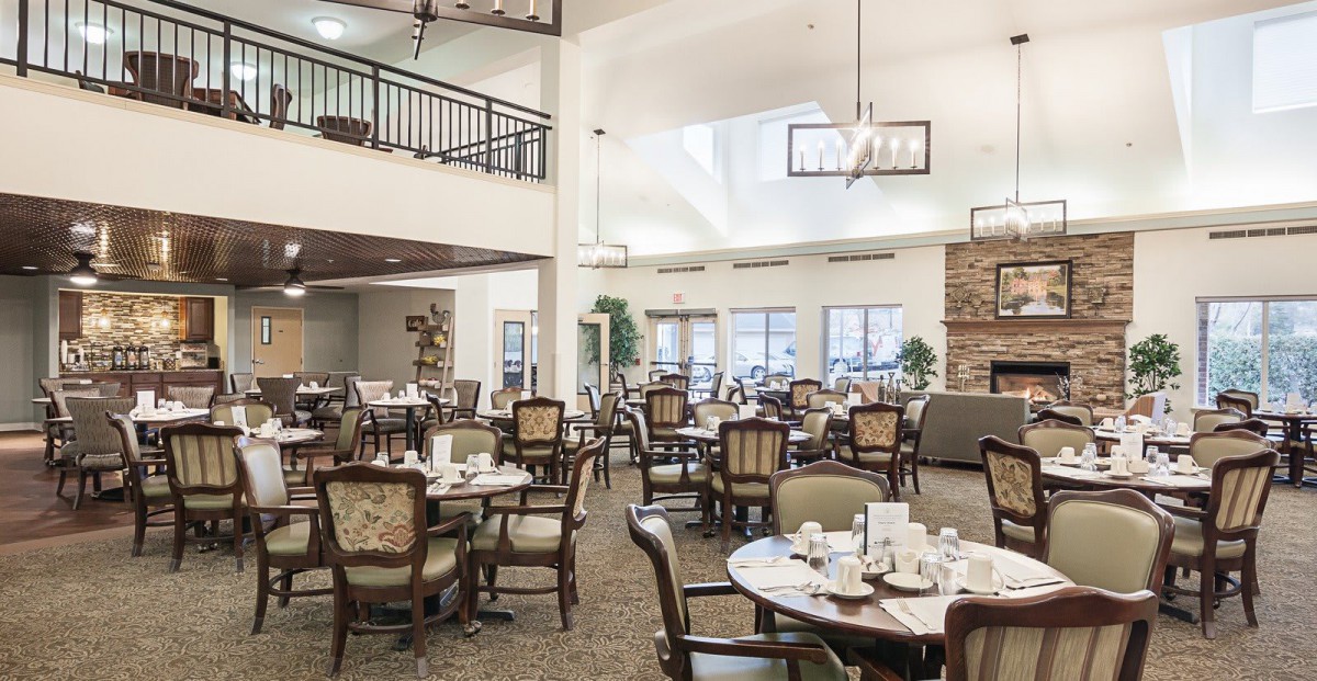 A spacious dining room with elegantly set tables and chairs, illuminated by chandeliers and natural light.
