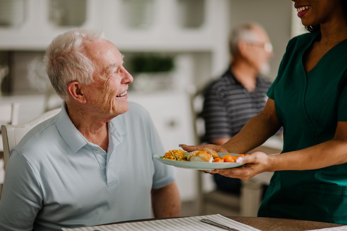 A caregiver serves a meal to an elderly man in a senior living facility.