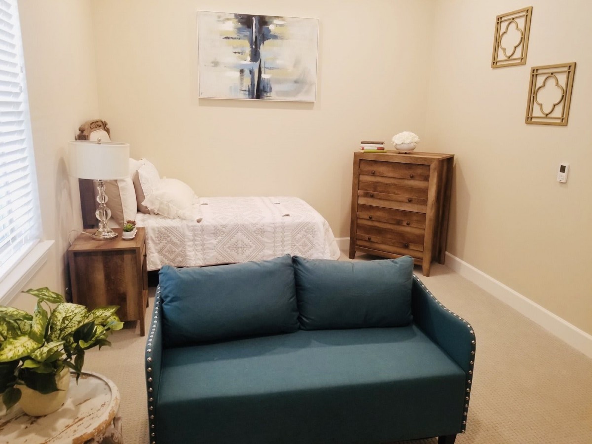 A cozy bedroom with blue sofa, bed, wooden dresser, and wall art.