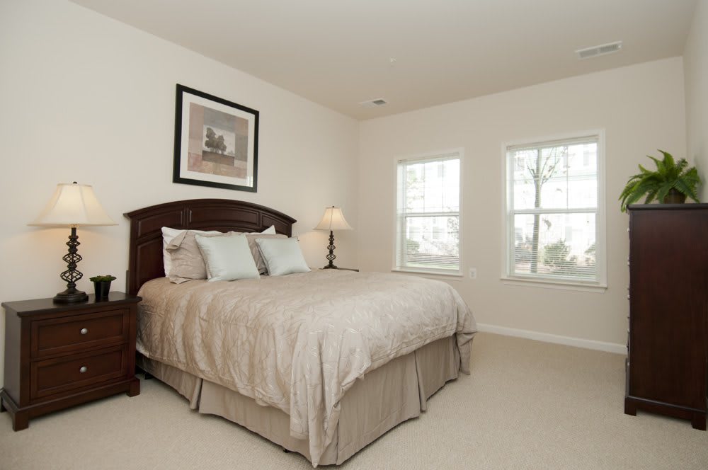 A cozy bedroom featuring a large bed with neutral bedding, flanked by a bedside lamp and artwork on the wall. Natural light streams in through two windows.