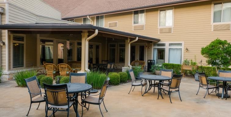 An outdoor patio area with tables and chairs, surrounded by a well-maintained garden and building exterior.