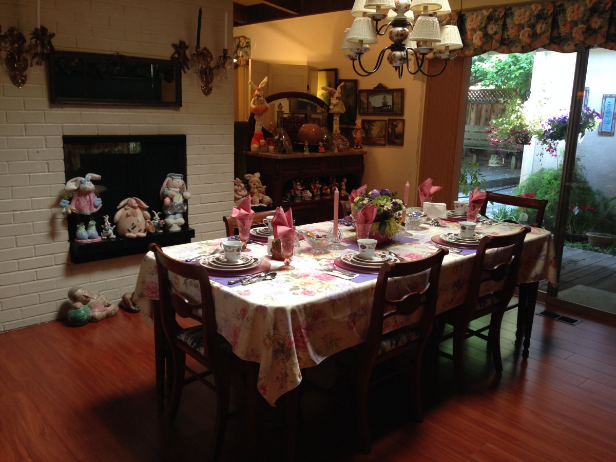 A warmly decorated dining room with a table set for a meal, featuring a chandelier and large window providing natural light.