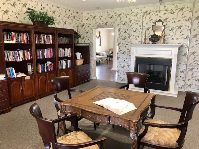 A cozy library room with a wooden table, chairs, bookshelf, and a fireplace.