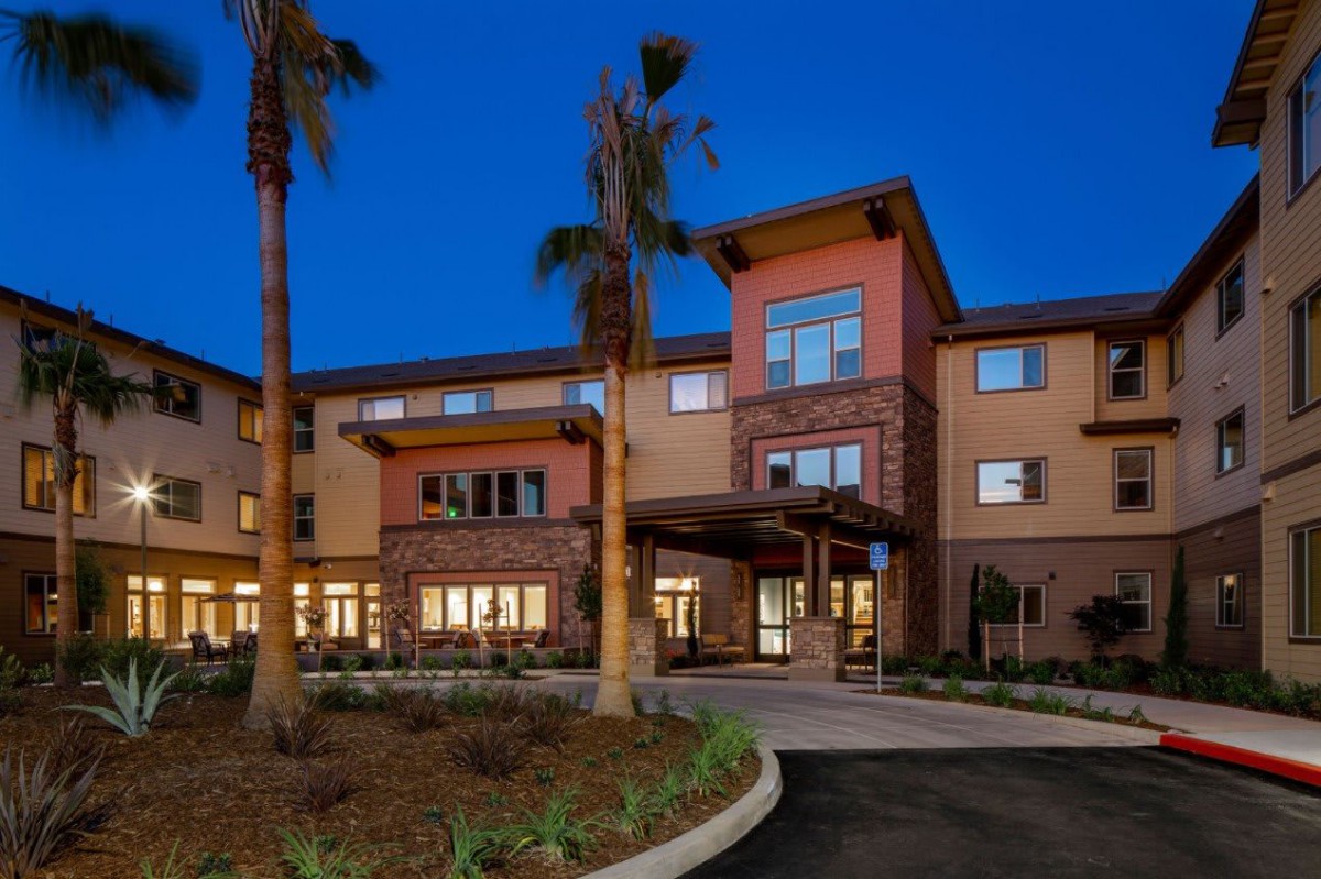 A modern residential building with palm trees, illuminated in the evening.