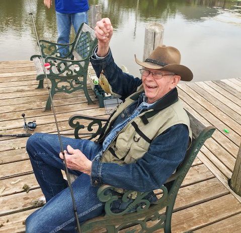 An elderly man sitting on a dock, holding up a small fish he caught, while wearing a cowboy hat.