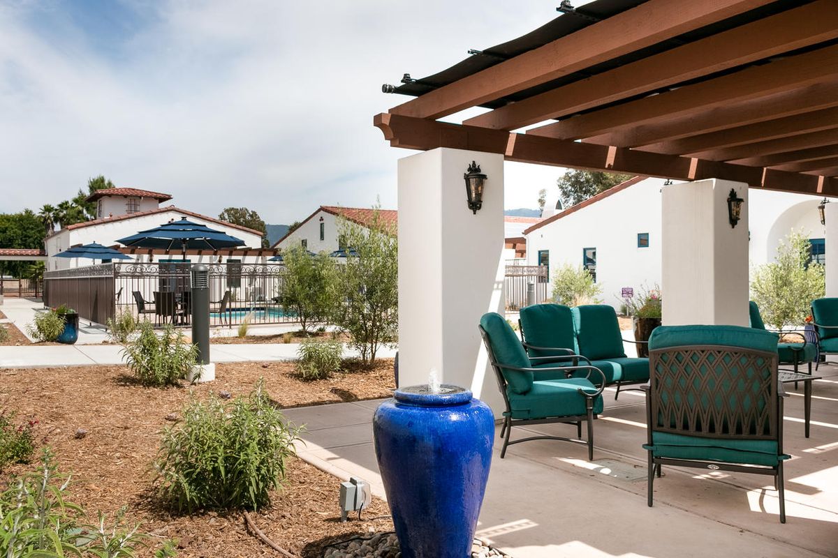 A serene outdoor patio with comfortable seating and a blue vase under a pergola, surrounded by well-maintained landscaping.