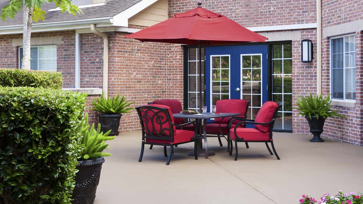 A cozy outdoor patio setup featuring red cushioned chairs and a matching umbrella, set against a brick wall with French doors.