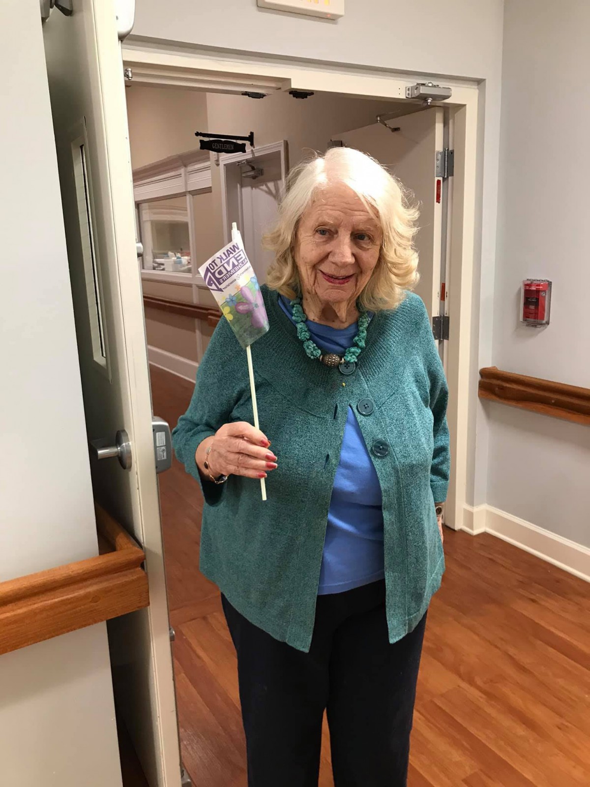 An elderly woman stands in a hallway holding a flower and smiling.