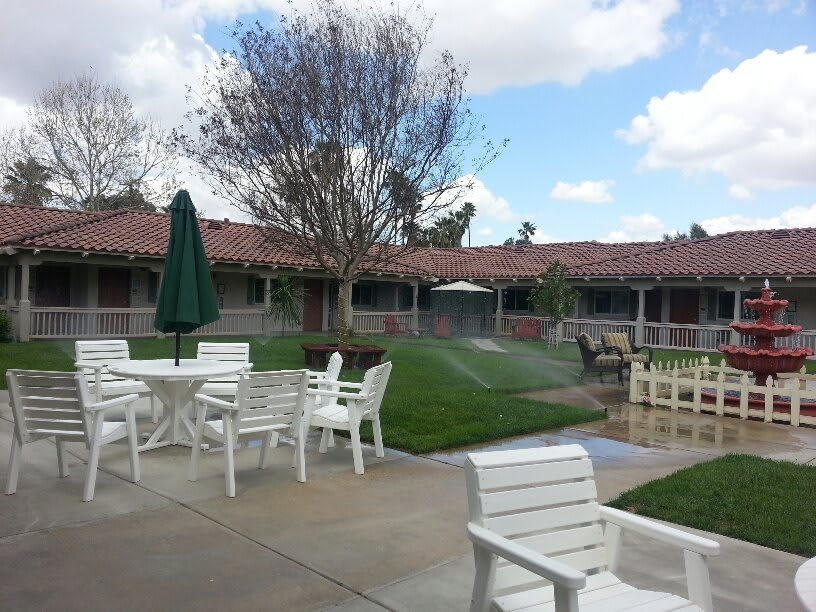 A peaceful patio area with white furniture overlooking a well-maintained lawn and trees under a partially cloudy sky.