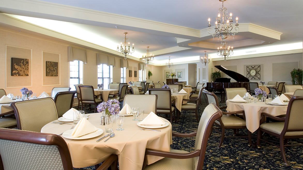 An elegant dining room with neatly arranged tables, chandeliers, and a grand piano.