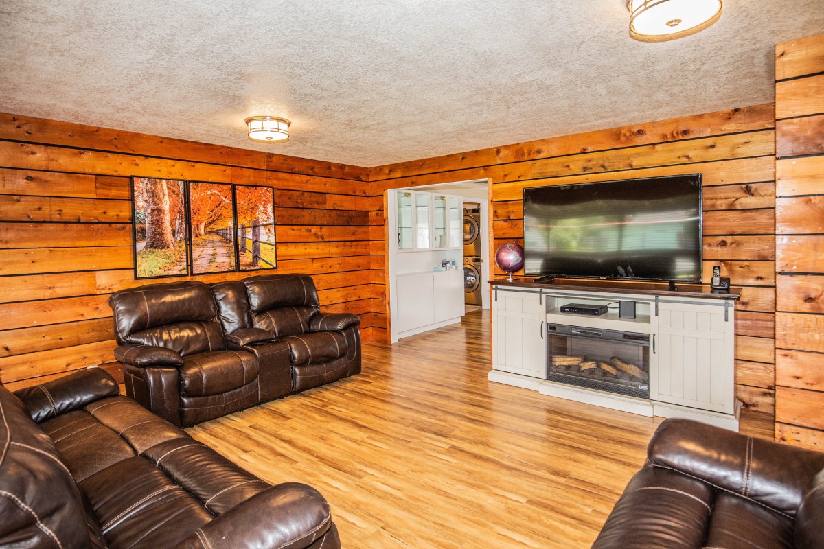 A cozy living room featuring leather couches, wood paneling, a large TV above a fireplace, and hardwood flooring.