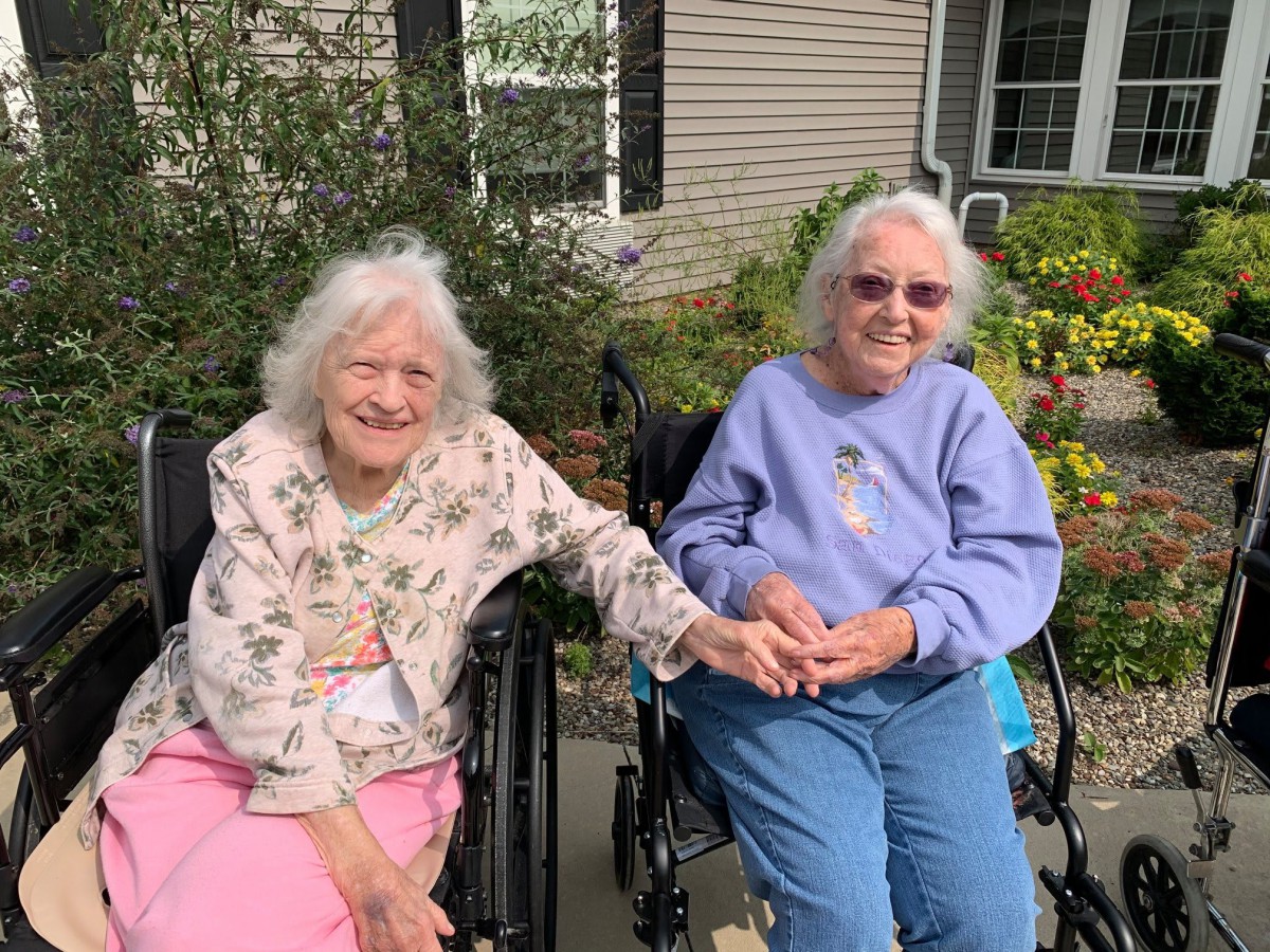 Two elderly women sitting in wheelchairs, holding hands and smiling outdoors.
