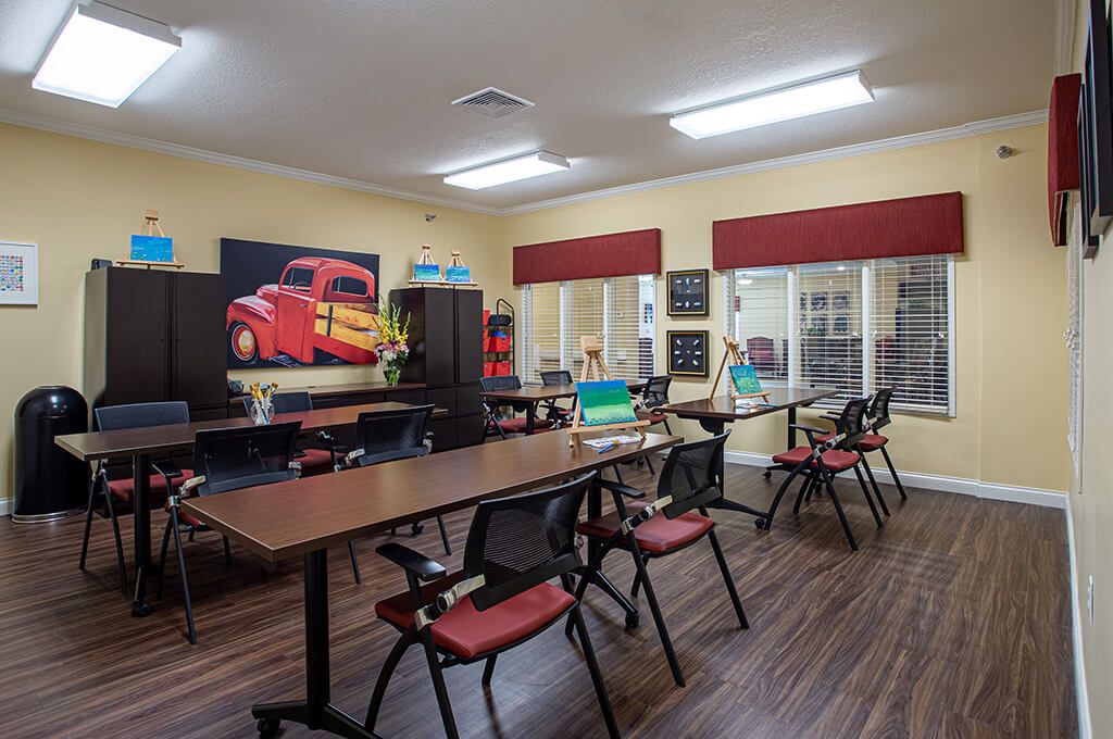 A classroom setting with tables and chairs, featuring artwork on the wall and large windows.