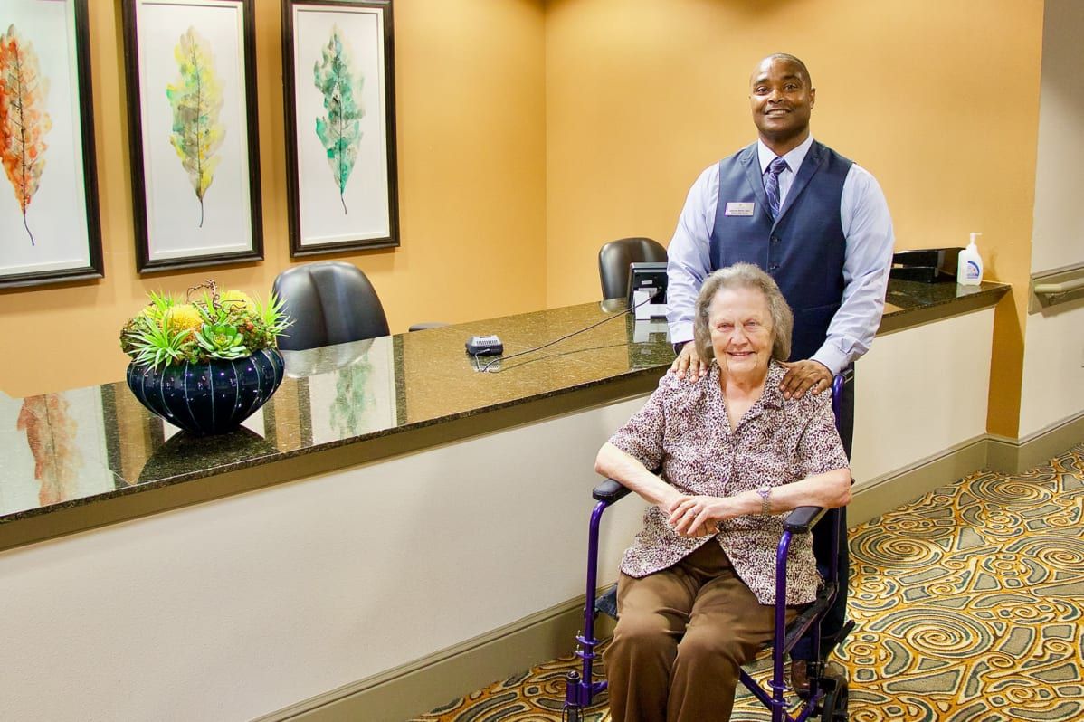 A caregiver standing behind an elderly woman in a wheelchair at a reception area.