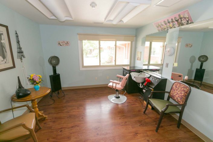 A cozy hair salon with wooden flooring and comfortable chairs, featuring a large window and mirror.