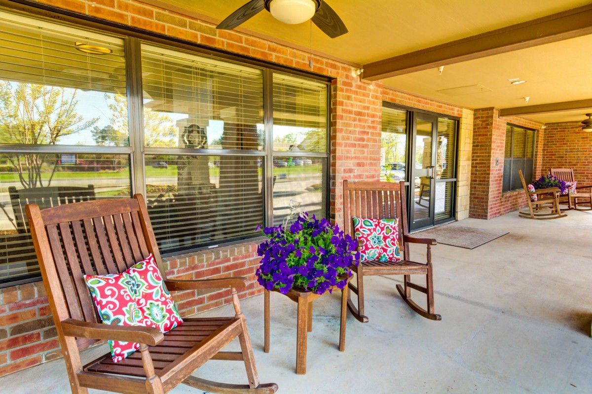 A cozy porch with rocking chairs, decorative pillows, and a small table with flowers in front of a brick house.