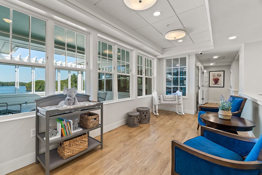 A bright hallway with wooden flooring and large windows, featuring seating and a bookshelf with decorative items.