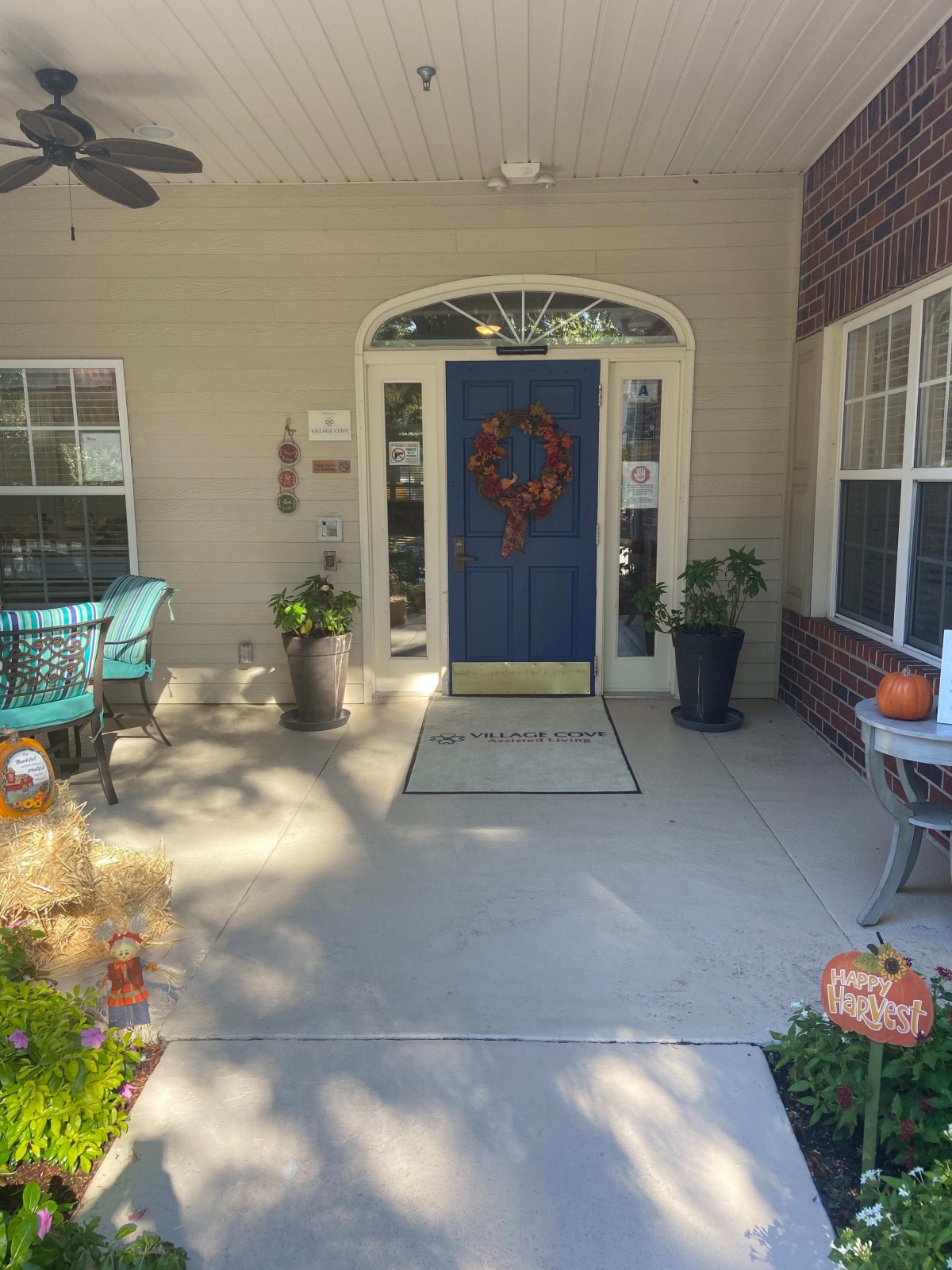 A cozy front porch with a blue door, adorned with a wreath and surrounded by fall decor, including plants and a pumpkin.