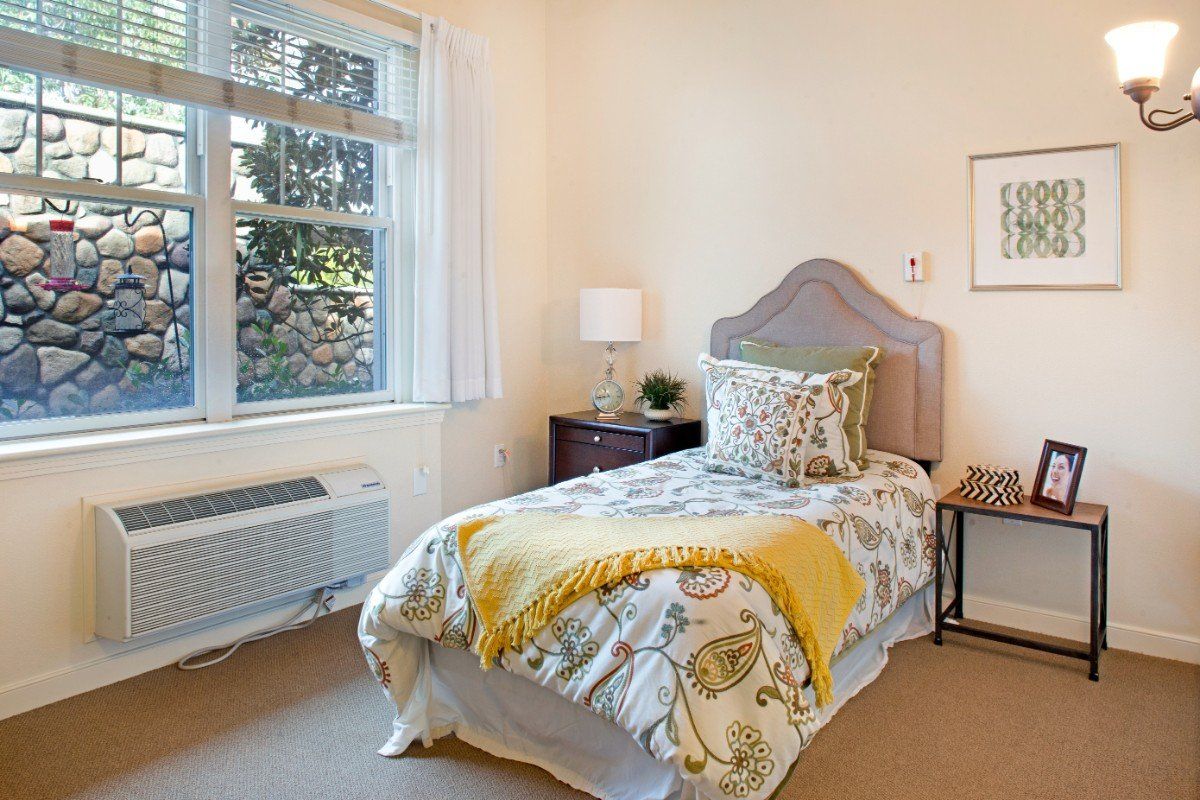A neatly arranged bedroom with a single bed, decorative pillows, and a nightstand with a lamp near the window.