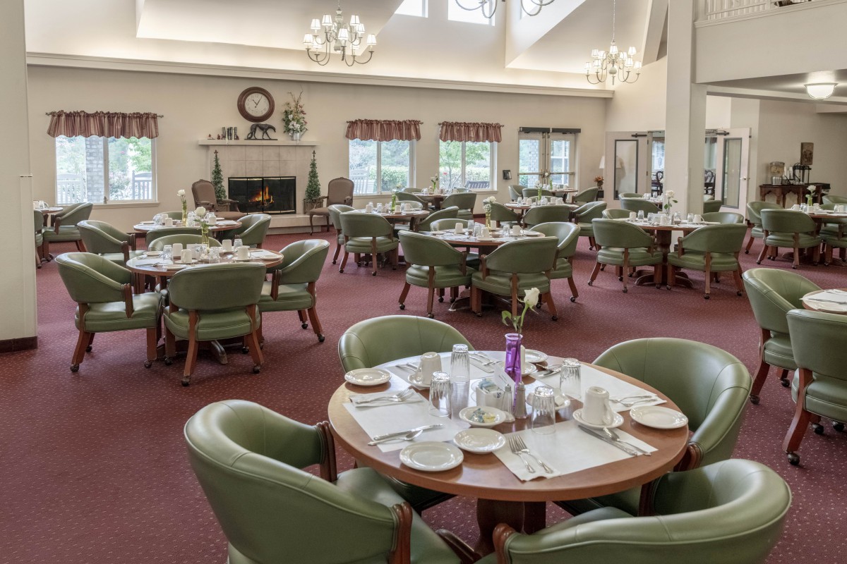 A spacious dining room with multiple tables set for a meal, featuring green chairs and large windows.