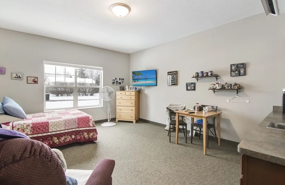 A cozy bedroom interior with a bed, dresser, table, and chairs, featuring wall decorations and a large window.