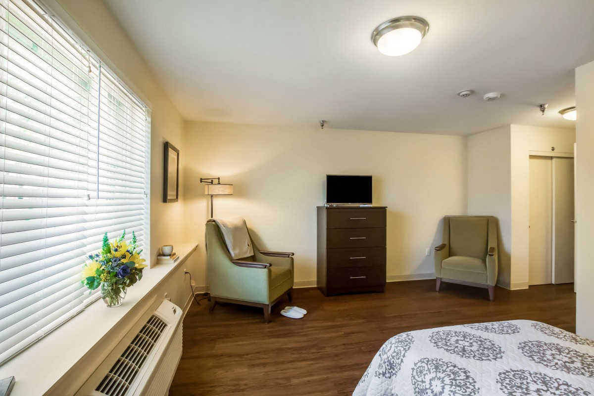 A cozy bedroom with a TV, armchairs, and a window letting in natural light.