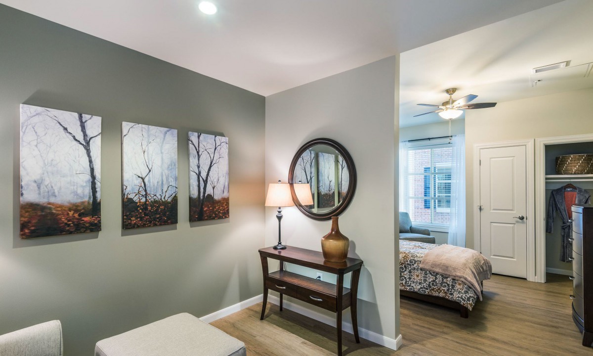 A cozy interior with a hallway leading into a bedroom, featuring wall art, a mirror, and a table lamp.