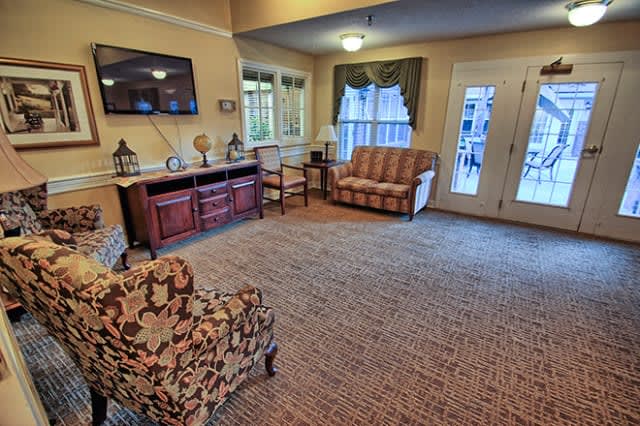 A cozy living room with classic furniture, including an armchair and sofa, carpeted floor, and natural light streaming through the windows.
