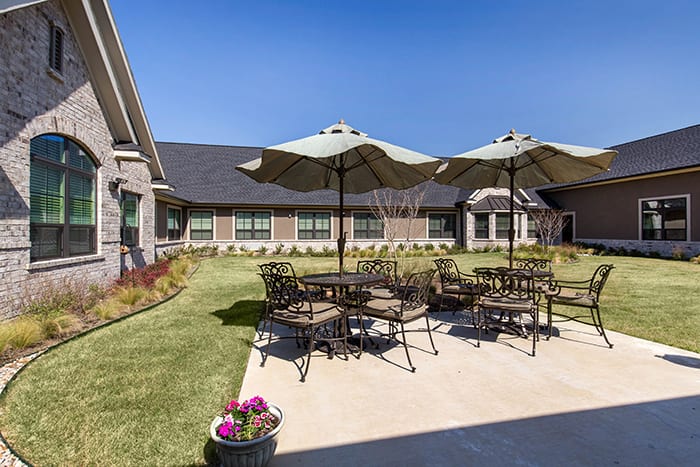 A sunny outdoor patio with tables, chairs, and umbrellas on a well-maintained lawn in front of a building.