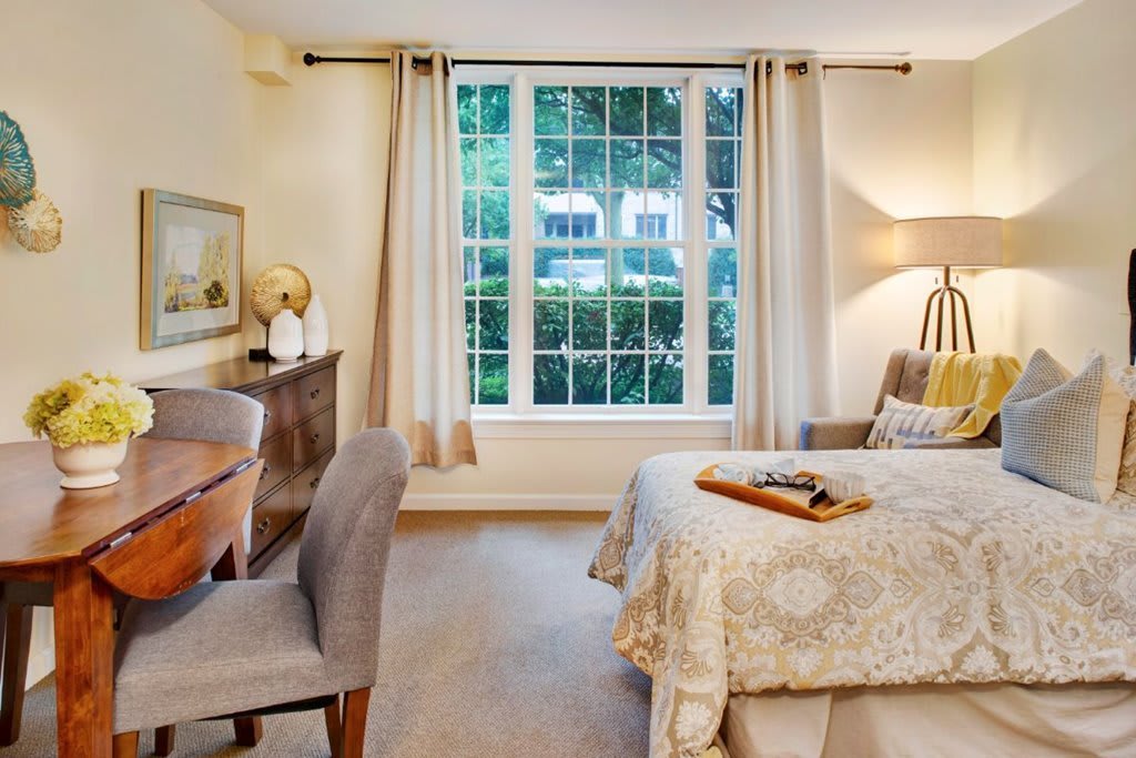 A cozy bedroom with a large window letting in natural light, featuring a comfortable bed, a desk with a chair, and stylish decor.
