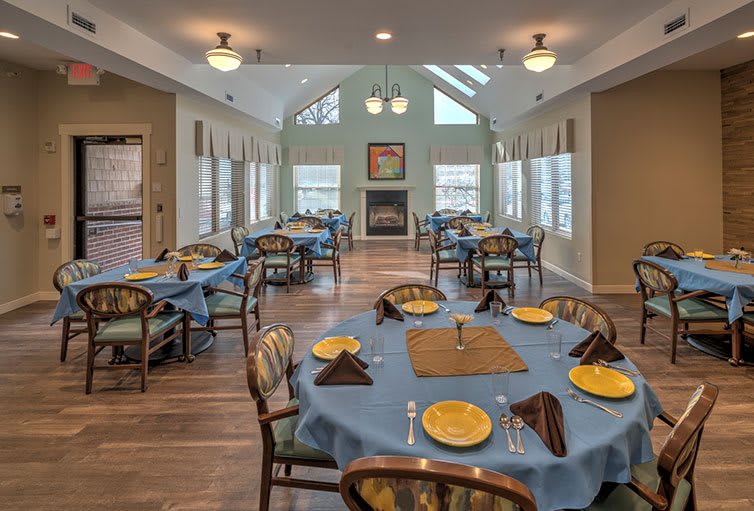 A cozy dining room with tables set for a meal, featuring blue tablecloths and ample natural light.