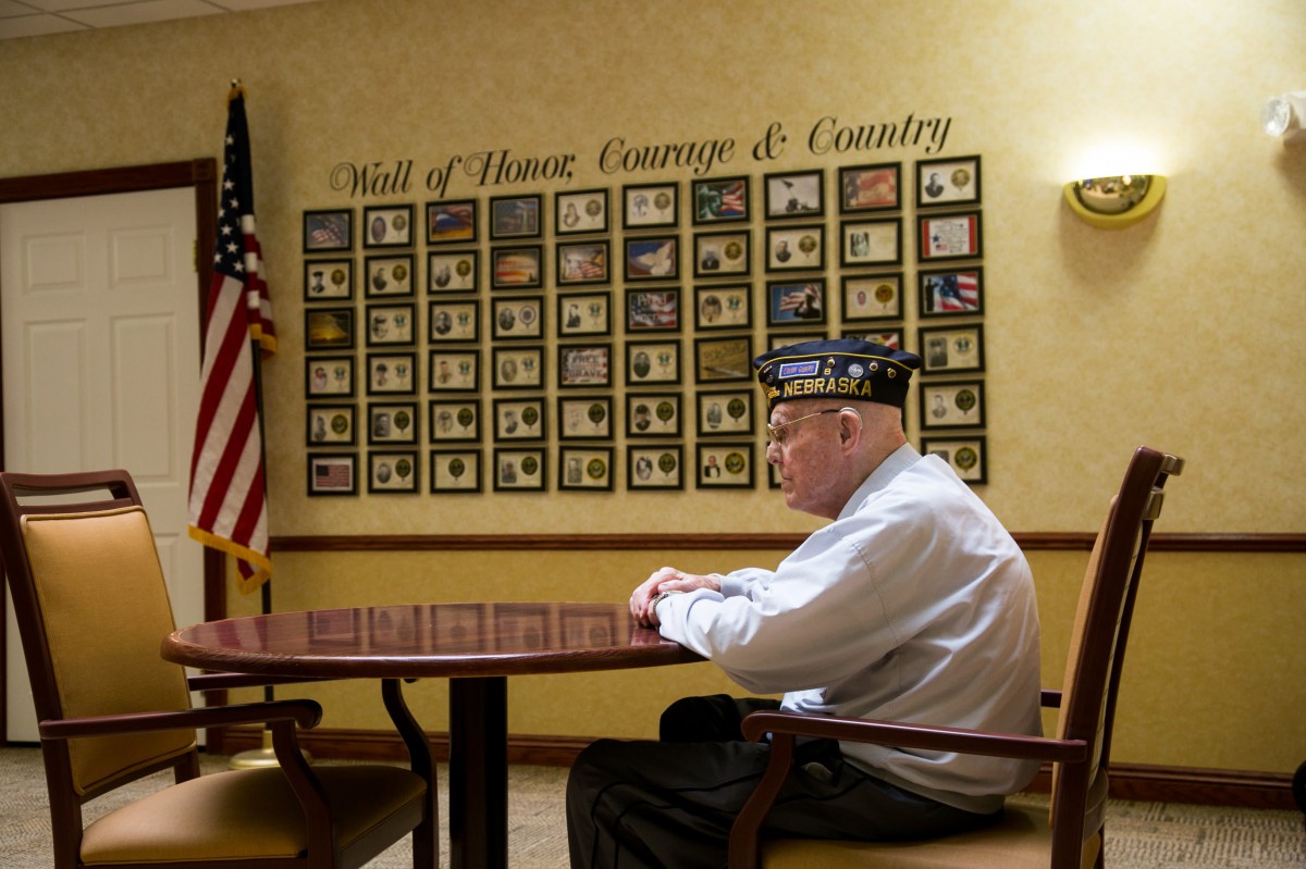 An elderly veteran sits in front of a wall displaying photographs and memorabilia, with an American flag nearby.