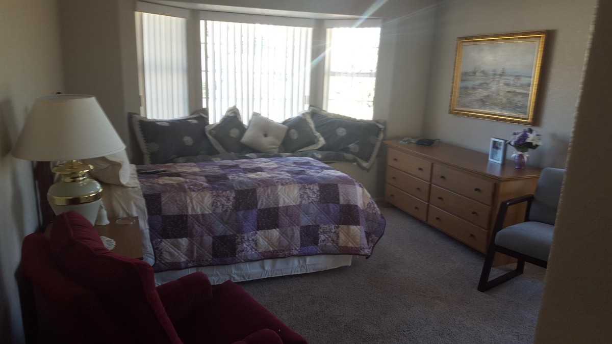 A cozy bedroom featuring a quilt-covered bed with multiple pillows, flanked by a window letting in natural sunlight. A dresser and soft carpet complete the decor.