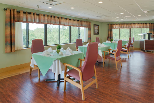 A well-lit dining area featuring neatly arranged tables with green tablecloths and comfortable chairs, surrounded by large windows.
