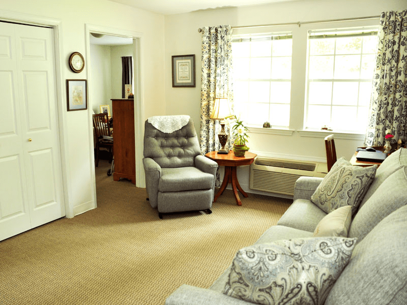 A cozy living room with a comfortable armchair and sofa arranged around a small table, well-lit by natural light through large windows.
