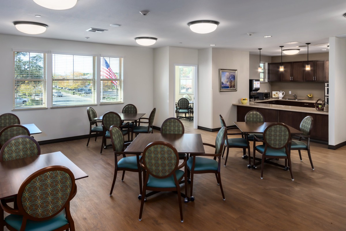 A cozy dining area with several tables and chairs, adjacent to a kitchen space, featuring large windows and overhead lighting.