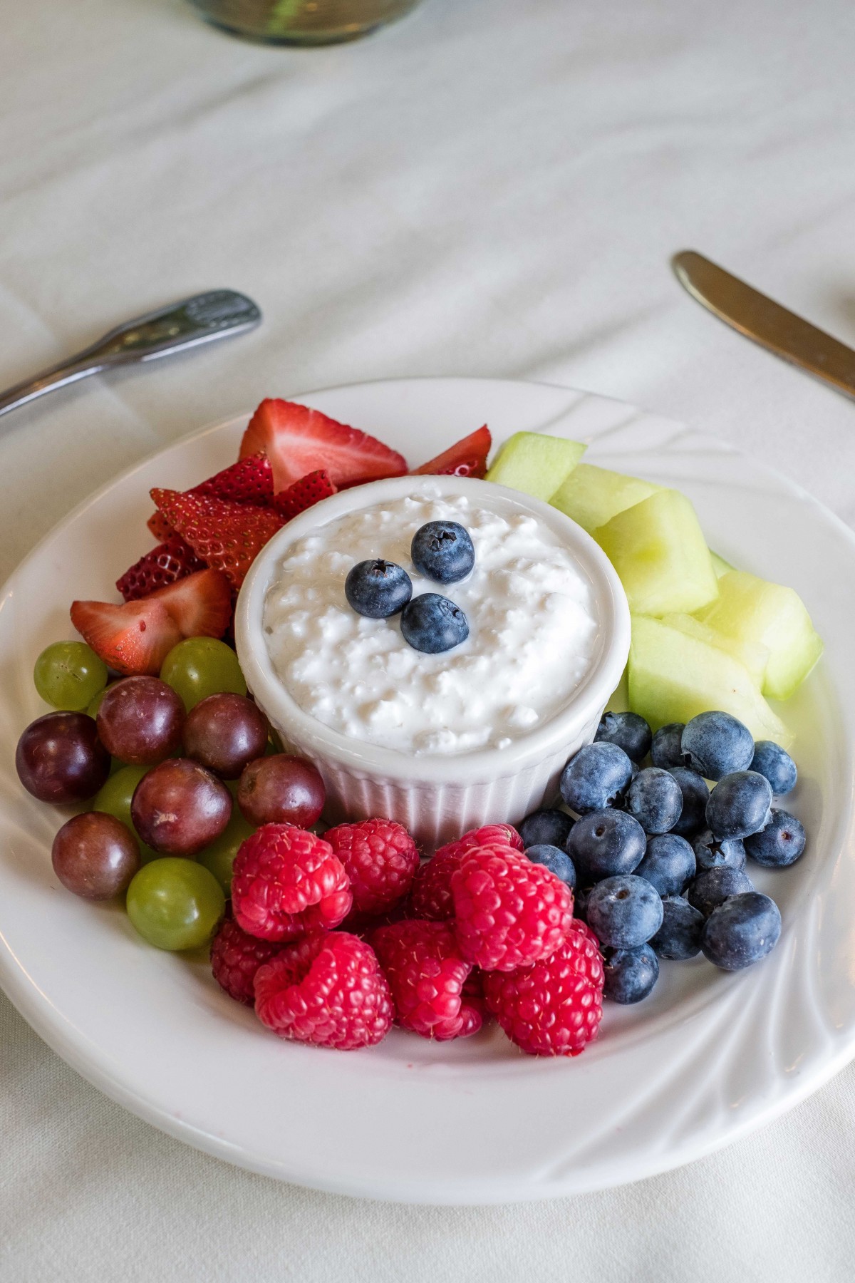 A healthy breakfast plate featuring yogurt surrounded by strawberries, blueberries, raspberries, grapes, and melon.