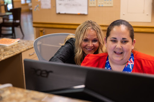 Two women are working together at a computer in an office setting, sharing a moment of joy.