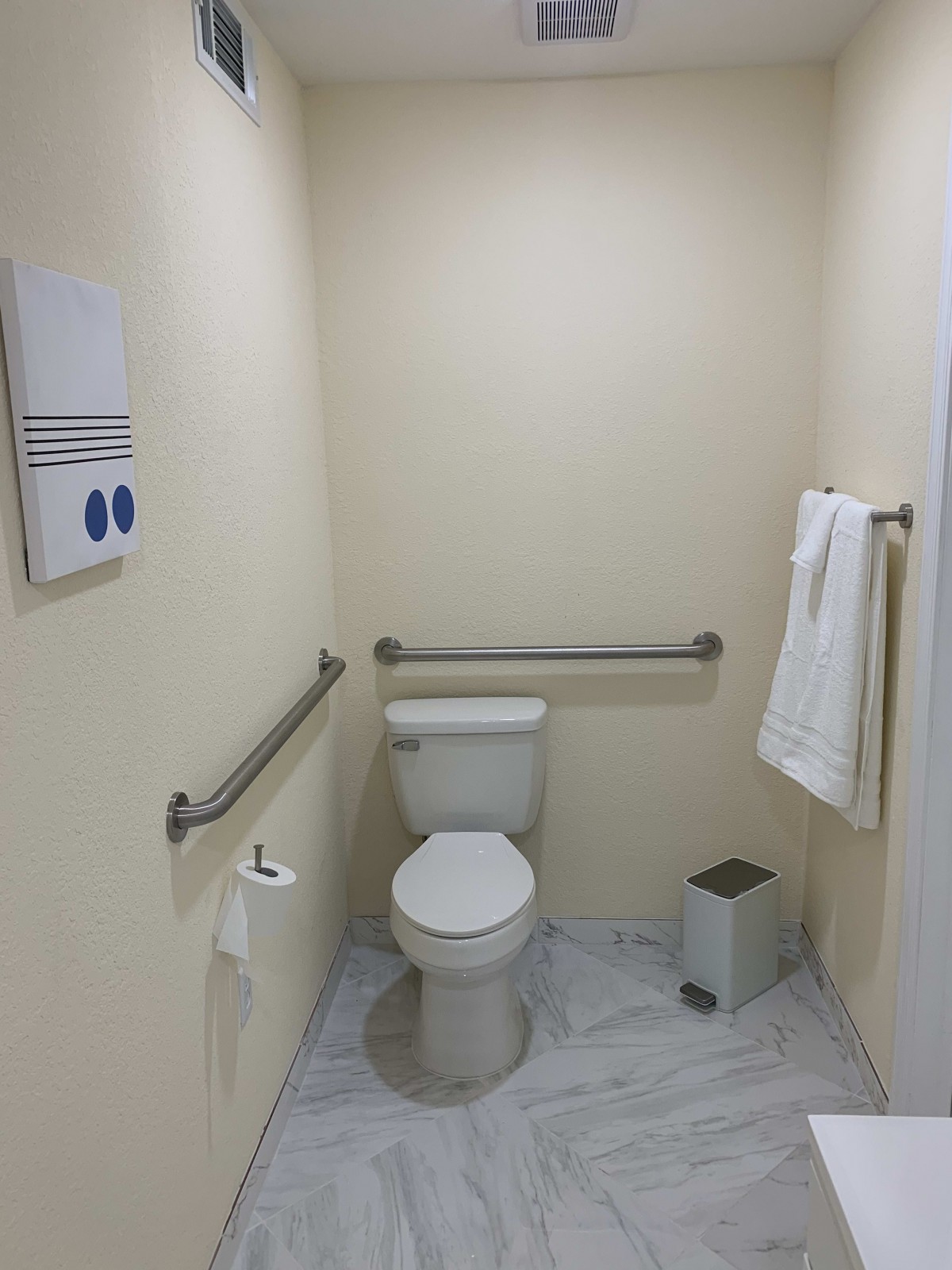A clean and simple bathroom featuring a toilet with handrails, towel rack, and waste bin.