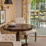 A cozy dining area with a round wooden table and upholstered chairs, featuring wall decor and curtains, overlooking a sunlit patio.