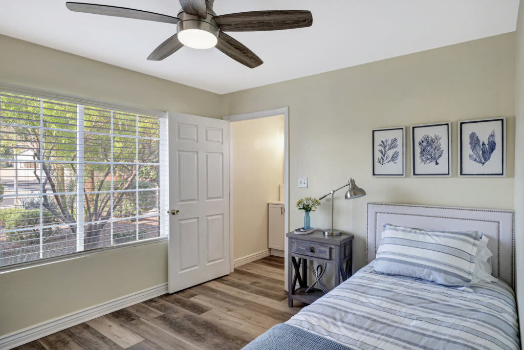 A cozy bedroom with a bed, side table, and ceiling fan, featuring natural light from a large window.