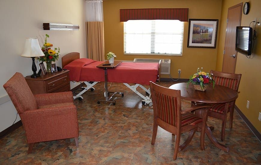 A well-furnished hospital room with a bed, table, chairs, and decorative flowers.