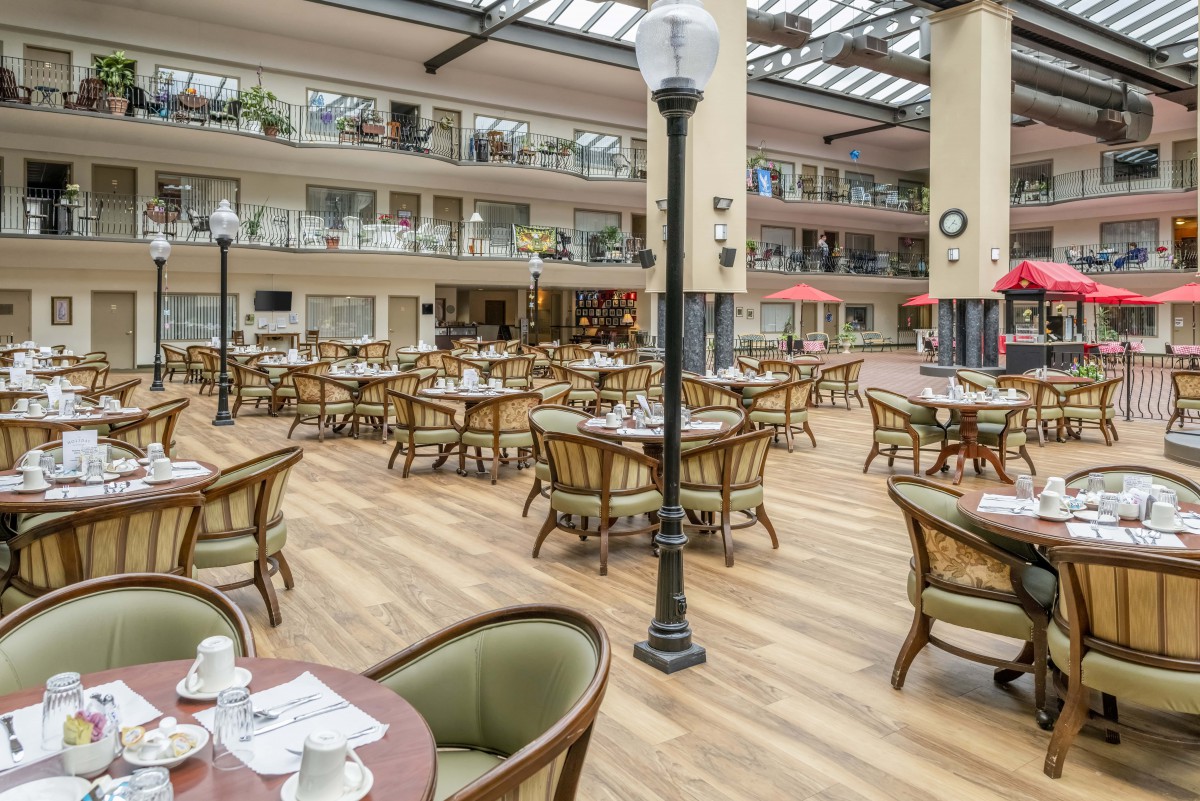 A spacious indoor dining area with multiple tables and chairs, featuring a glass canopy roof and decorative lighting.