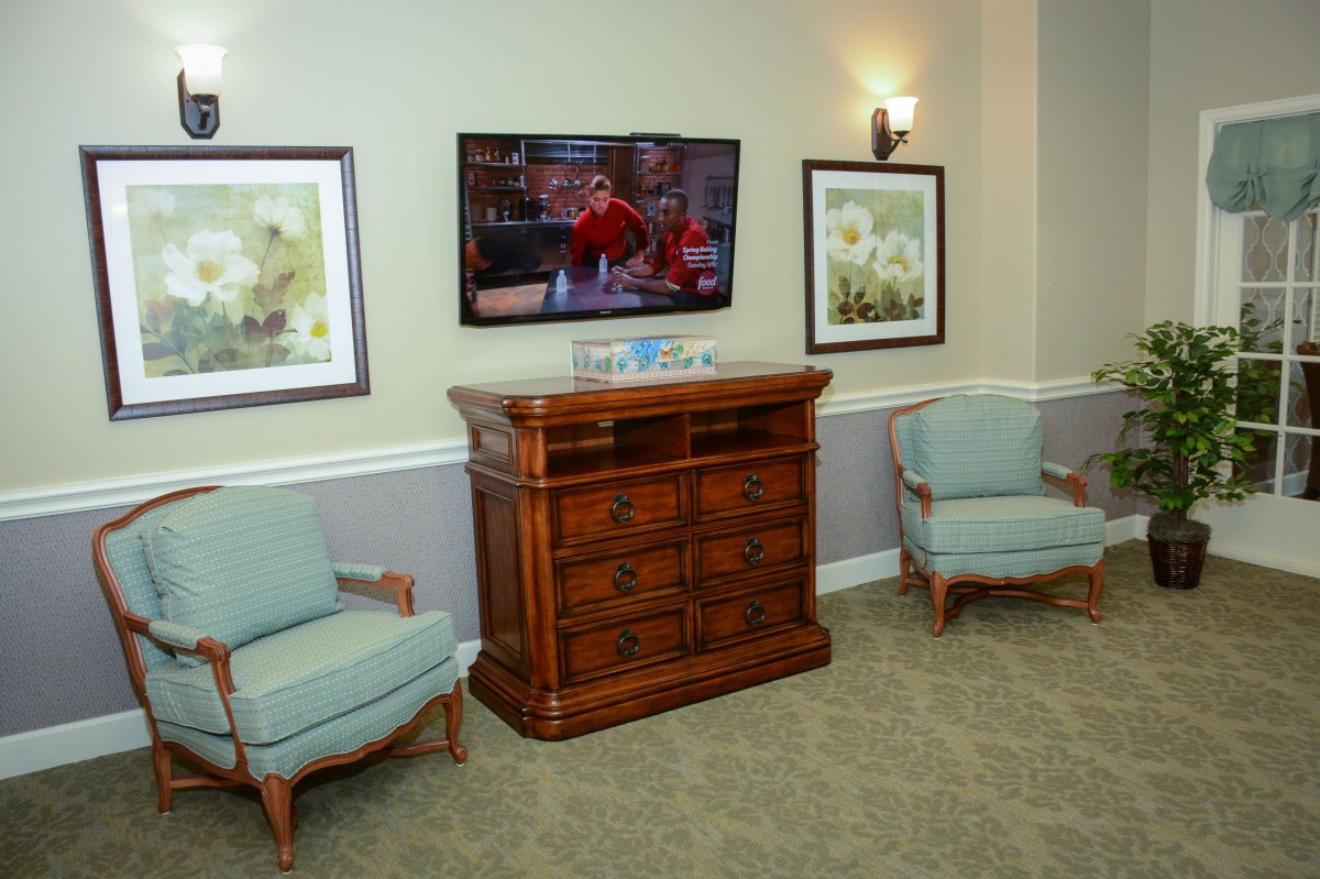 A cozy living room with armchairs and a television mounted above a wooden cabinet, flanked by framed paintings.