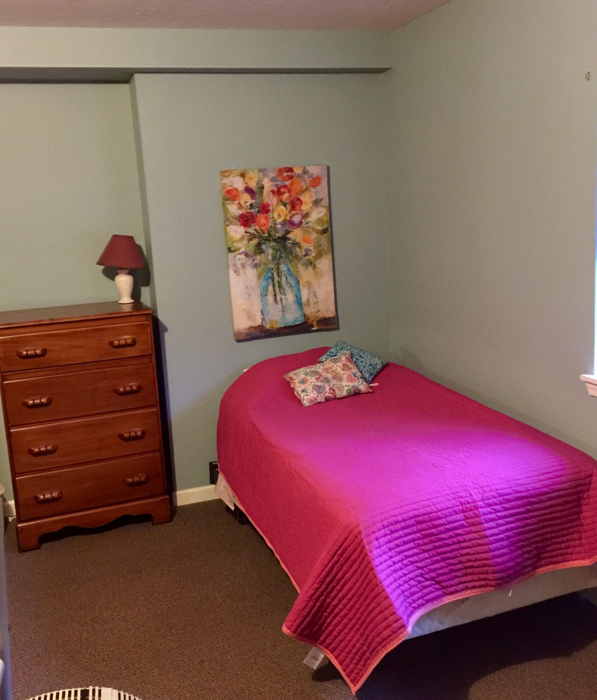 A cozy bedroom with a single bed adorned with a pink bedspread, next to a wooden dresser and floral artwork on the wall.