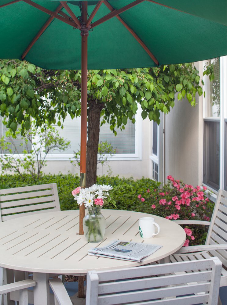A serene patio setup with a round table and chairs under a green umbrella, adorned with a vase of flowers.