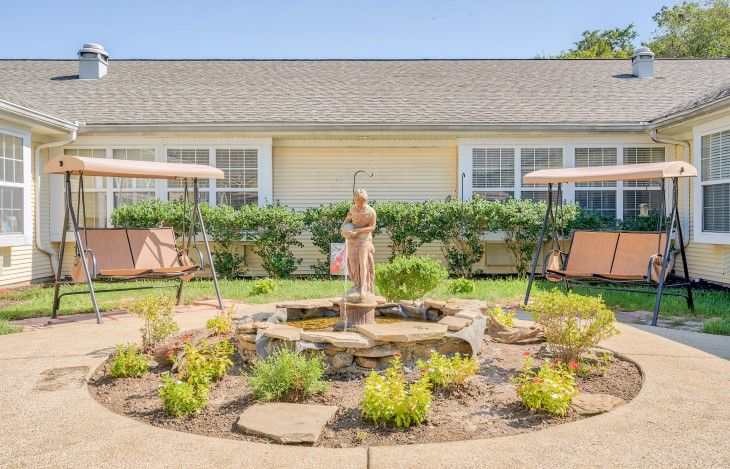 A picturesque courtyard featuring a central fountain with a sculpture, surrounded by swing benches and lush landscaping.