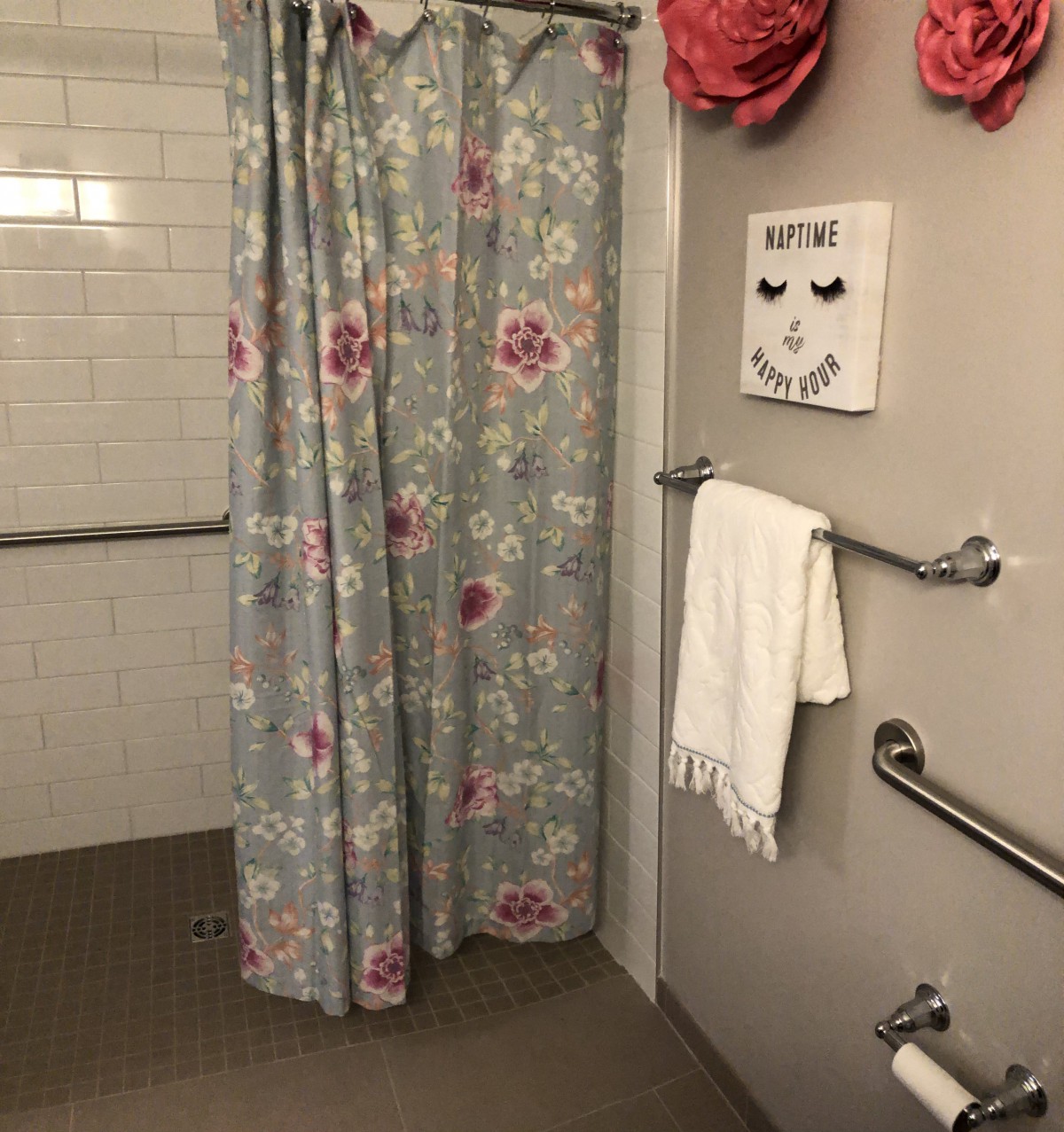 A bathroom with a floral shower curtain and handrails, featuring a towel rack and decorative wall art.